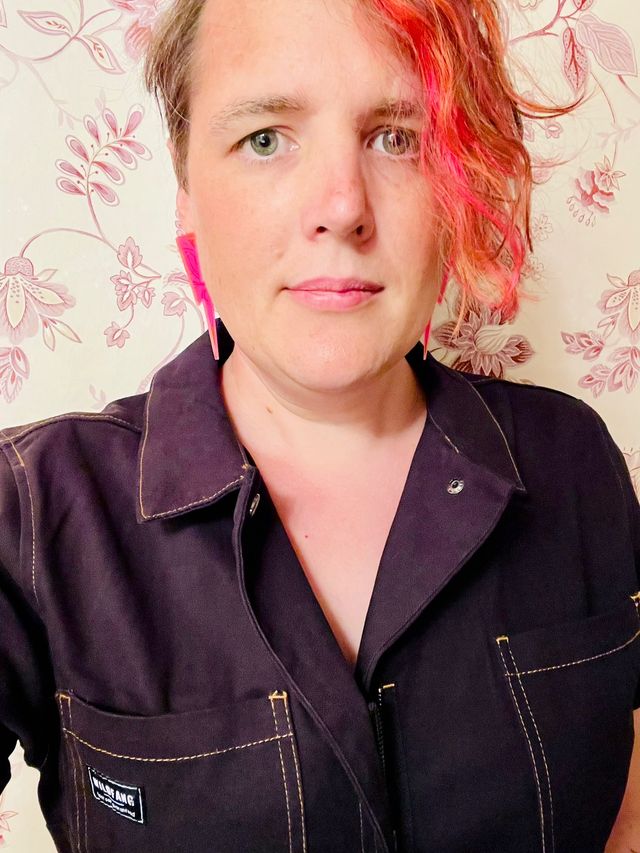 Miriam selfie in black coveralls
and neon pink lightning-bolt earrings,
with a lock of orange hair on one side.
