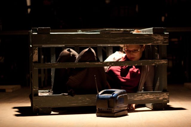 Julie crumpled in a wood crate watching a small TV through the slats